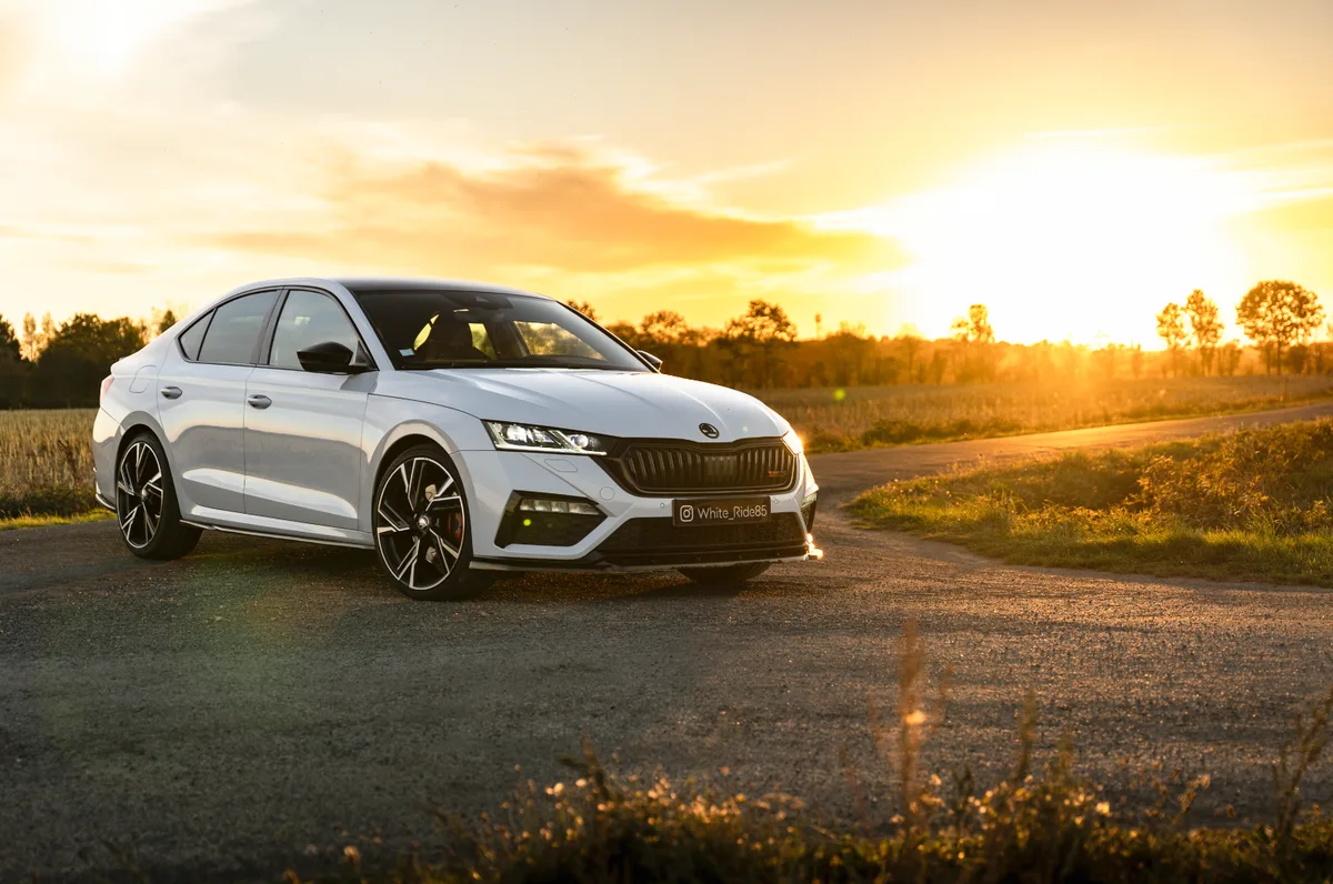 Berline Skoda blanche photographiée au coucher du soleil sur une route de campagne.