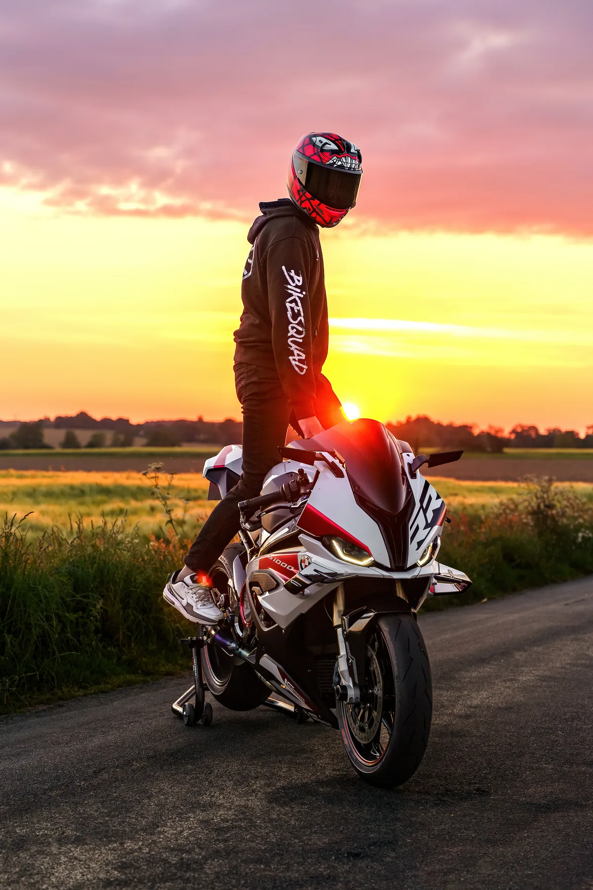 Un motard en casque rouge pose près de sa moto à la tombée du jour.