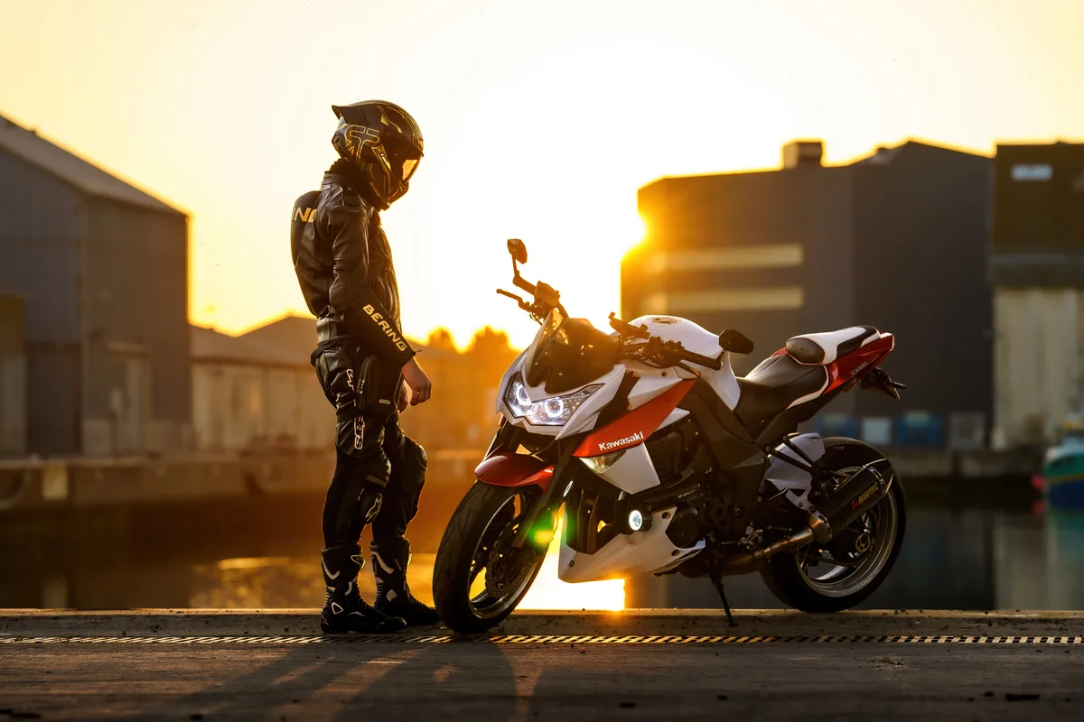 Un motard se tient près de sa moto rouge et blanche au soleil couchant.
