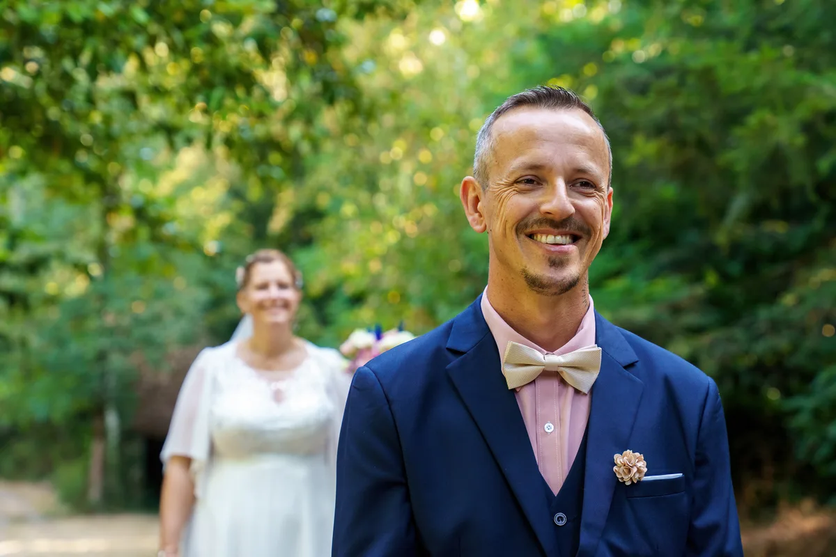 Portrait du marié souriant, avec la mariée en arrière-plan dans le jardin.