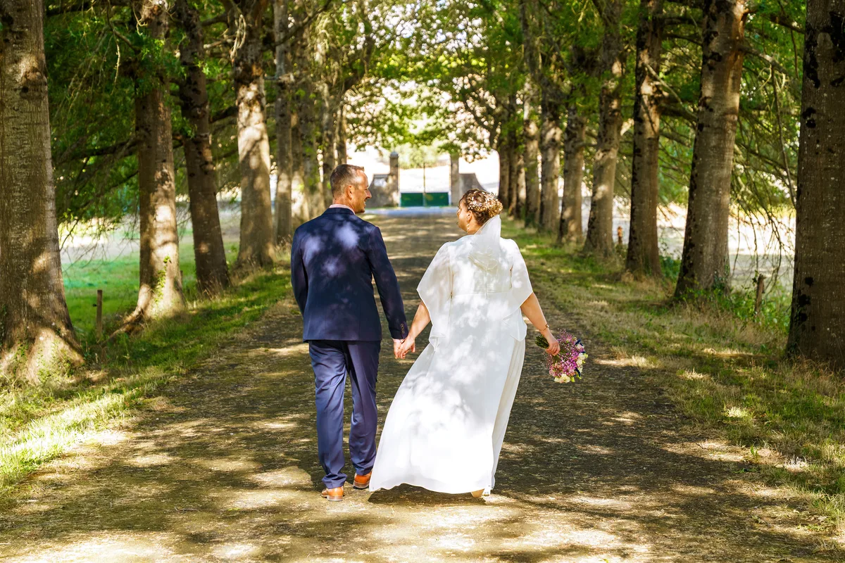 Les mariés s’éloignent main dans la main sur une allée bordée d’arbres.