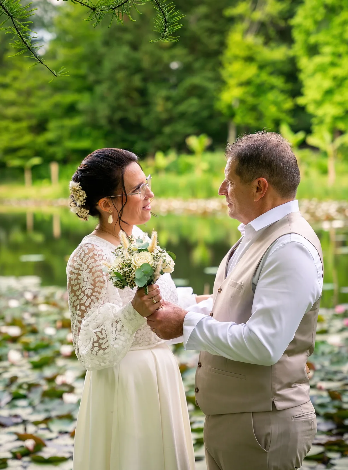 Les mariés se tiennent la main au bord d’un étang fleuri.