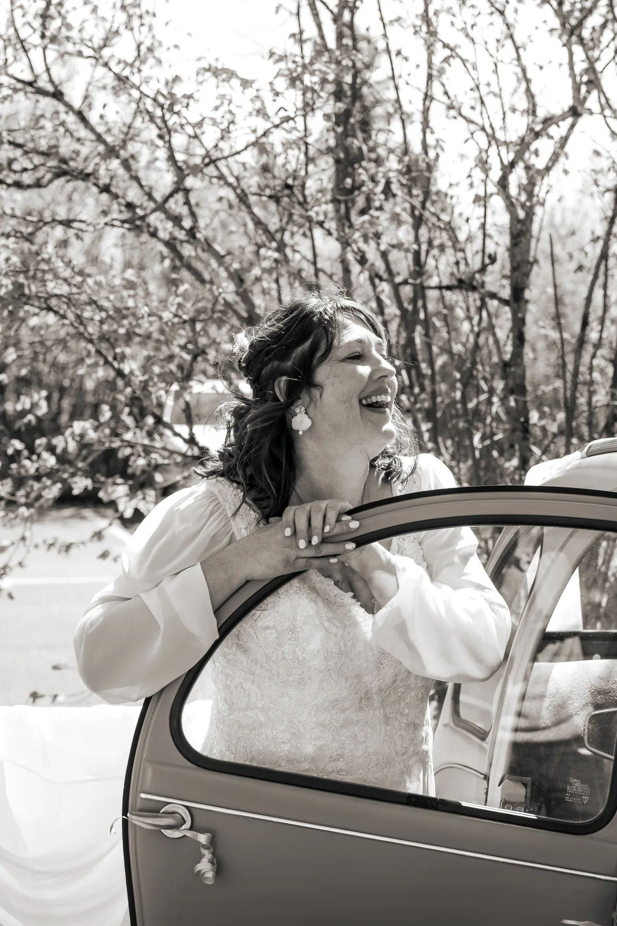 La mariée sourit depuis une voiture ancienne, photographiée en noir et blanc.